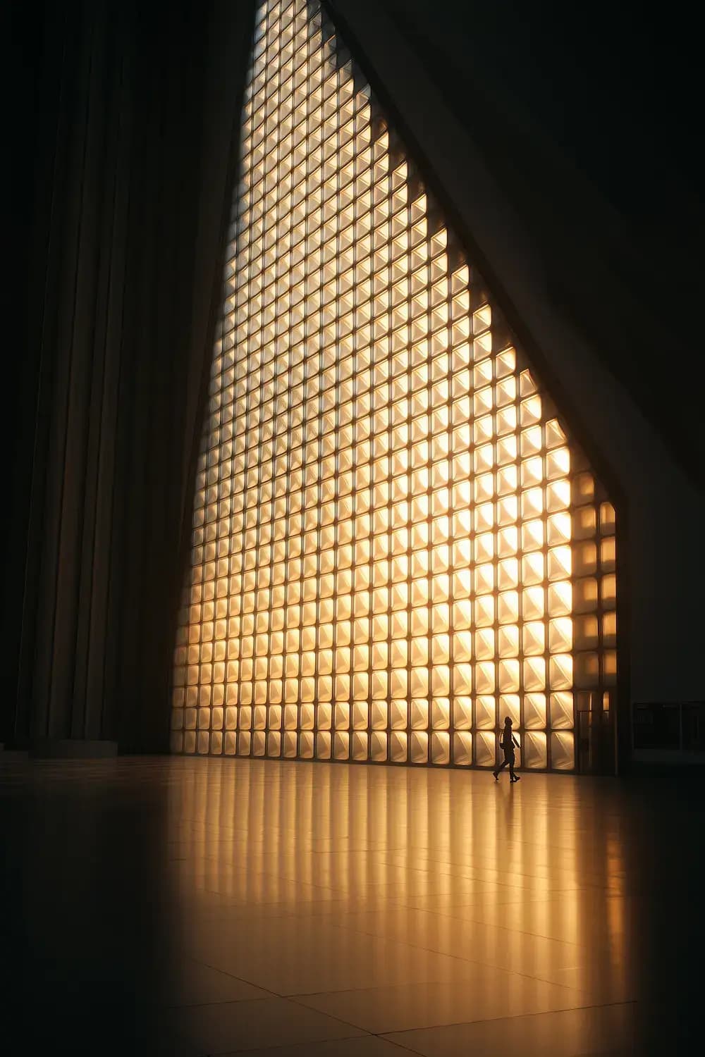 photography,triangular pattern translucent glass wall installation with black metal frame grid,warm golden backlight creating honeycomb shadows,silhouette of person walking,zen interior design style,amber glow atmospheric lighting,professional studio setup with two continuous led panels for backlight at 2800k,geometric repetitive patterns,high contrast interior shot,ultra sharp details,photorealistic quality,48K resolution,rich detailing